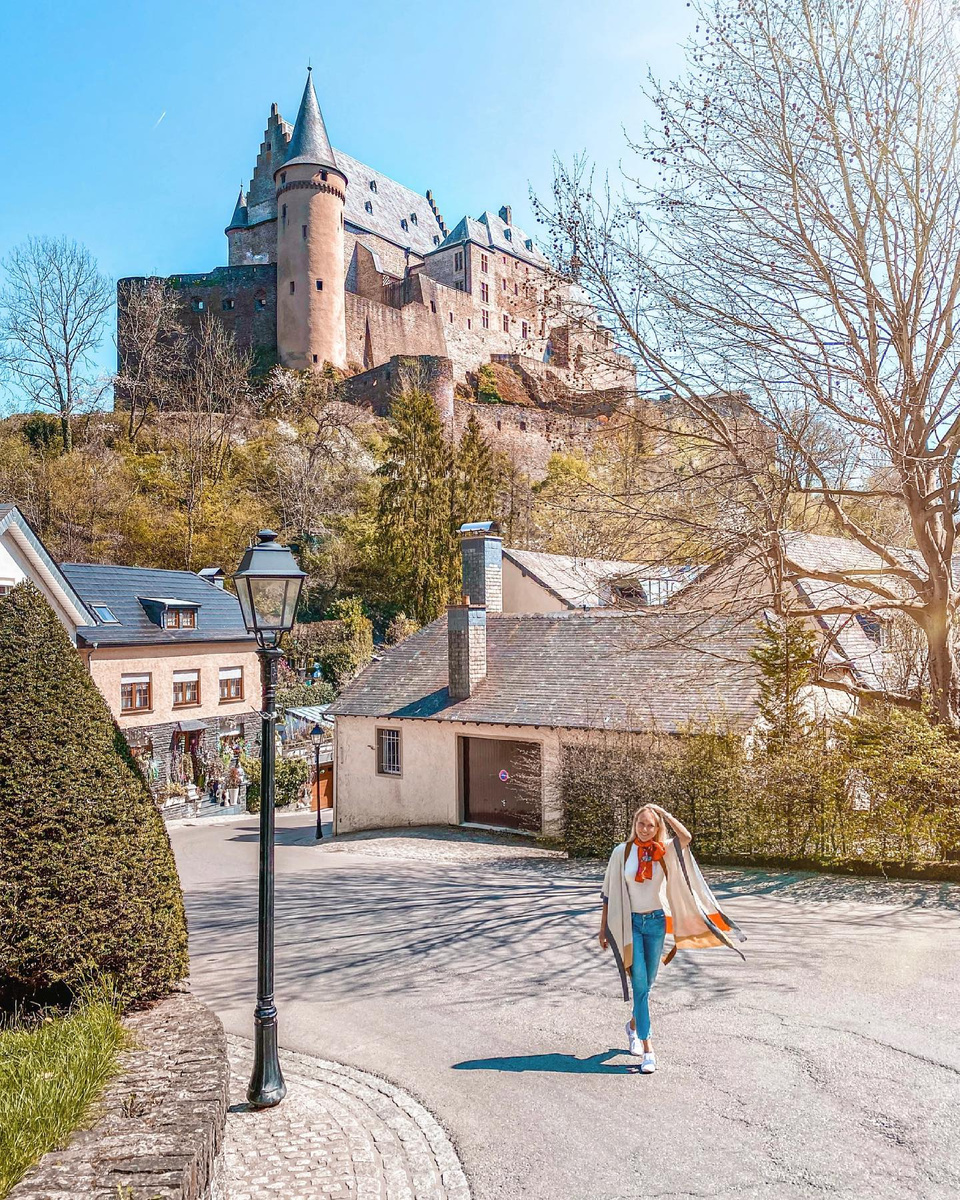 Olga walking up a village street beneath Vianden Castle in spring sunshine.