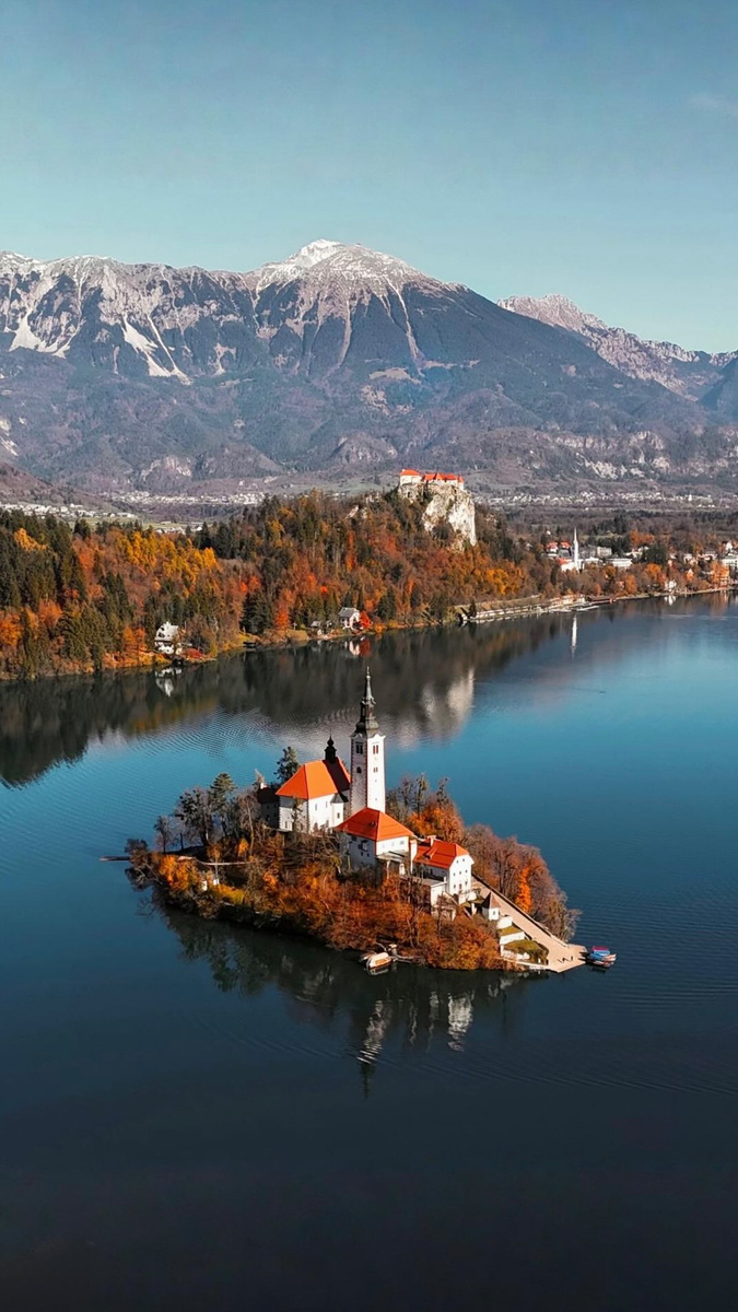 Bled Island and its church reflected in the lake beneath mountains and Bled Castle.