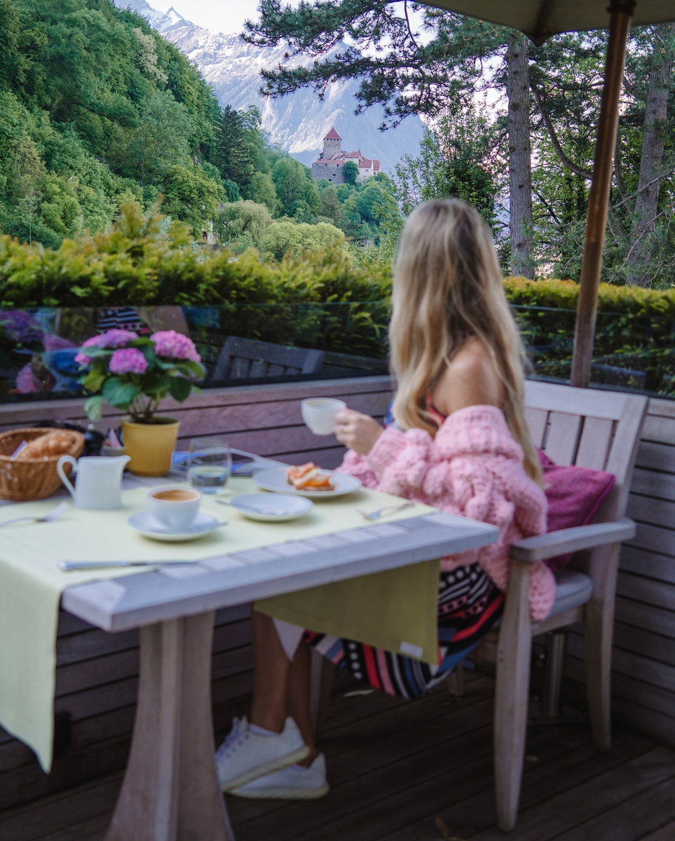 Olga at breakfast on the Sonnenhof terrace, with Vaduz Castle and the Alps beyond the trees.