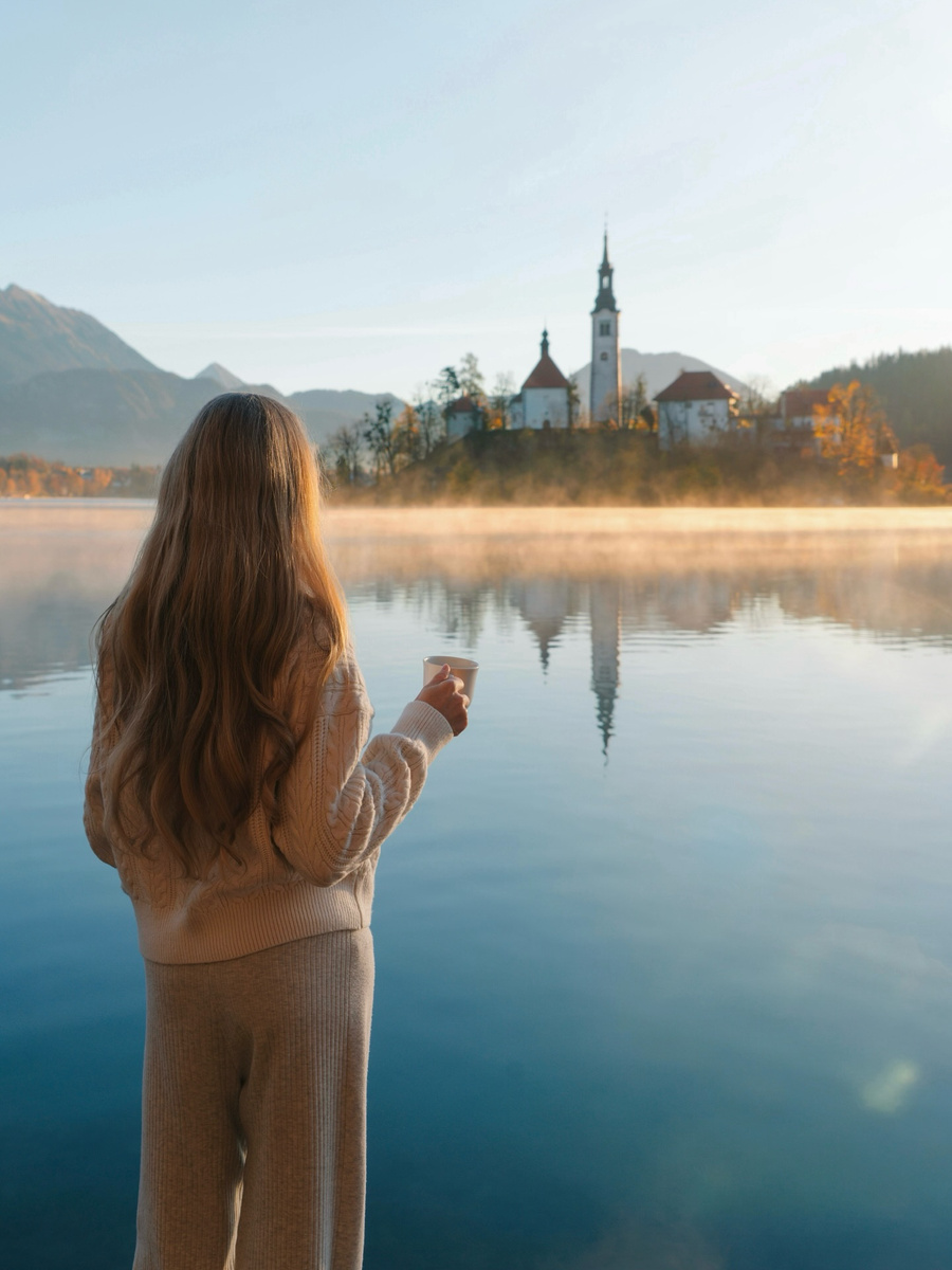 Olga holding a coffee cup beside Lake Bled at sunrise, with the island reflected in the water.