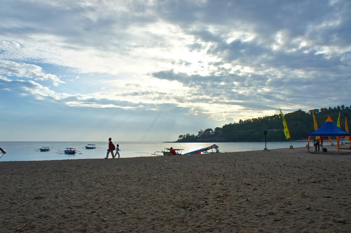People walking on Senggigi Beach beside moored boats beneath a clouded sky.