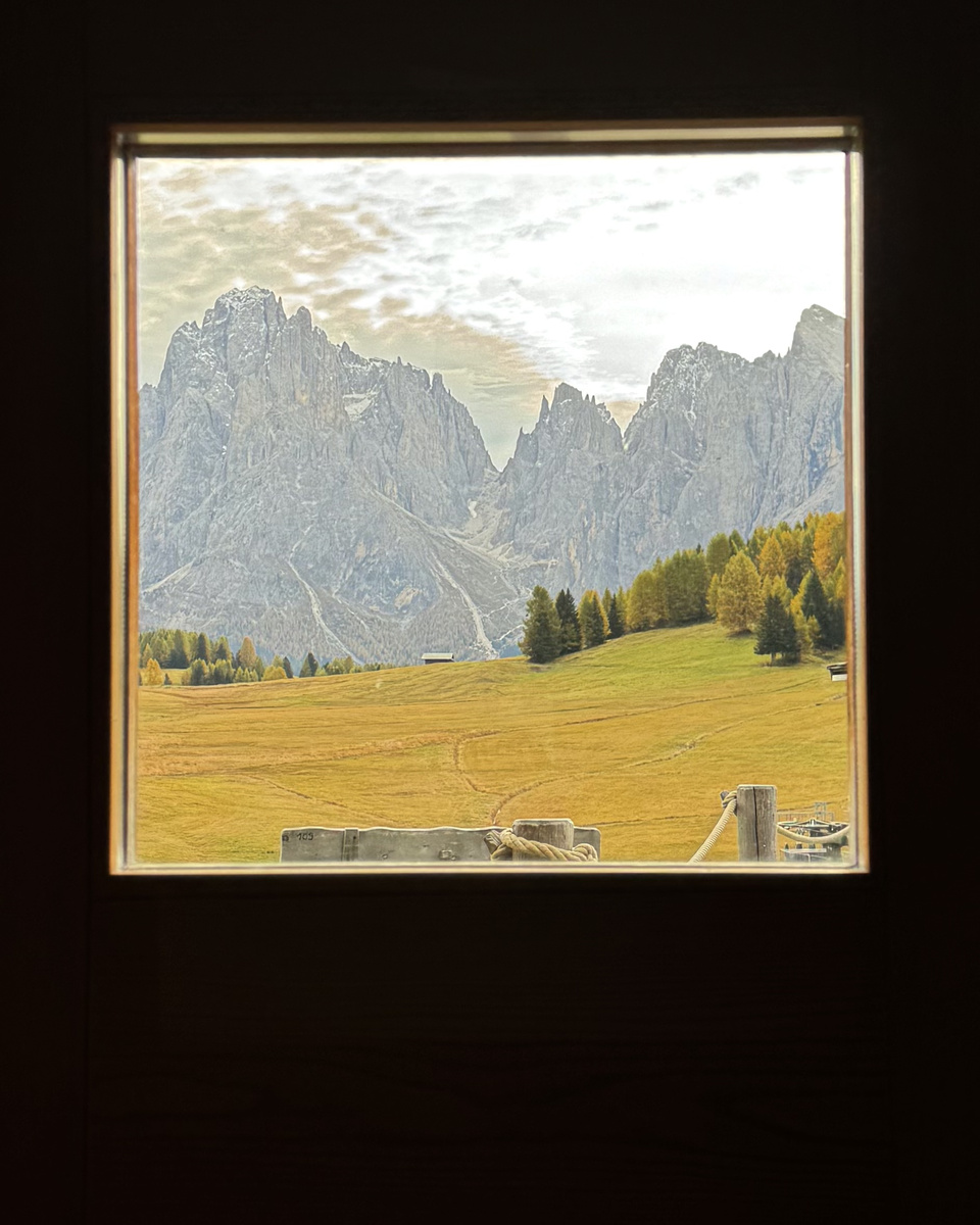 A wooden window frames a meadow, golden larches and the rocky Sassolungo group.