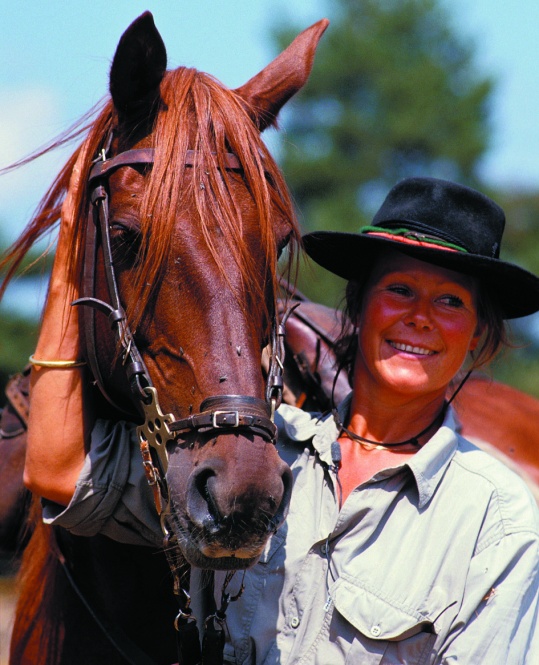 Anne Mariage smiles beside a bridled chestnut horse, wearing a dark riding hat.