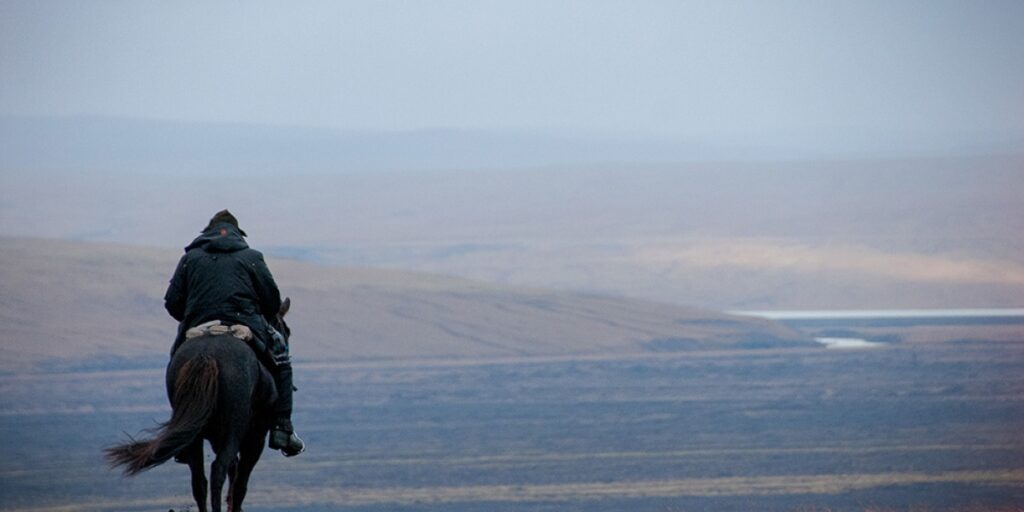 A lone rider on a dark horse crosses open country towards a distant lake.
