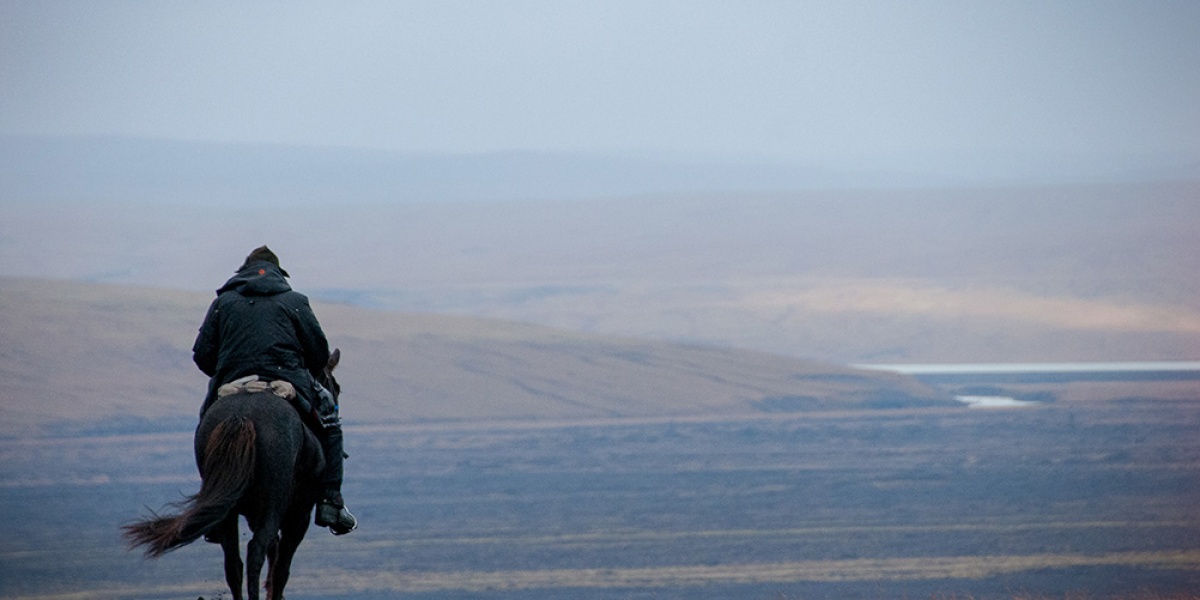 A lone rider on a dark horse crosses open country towards a distant lake.