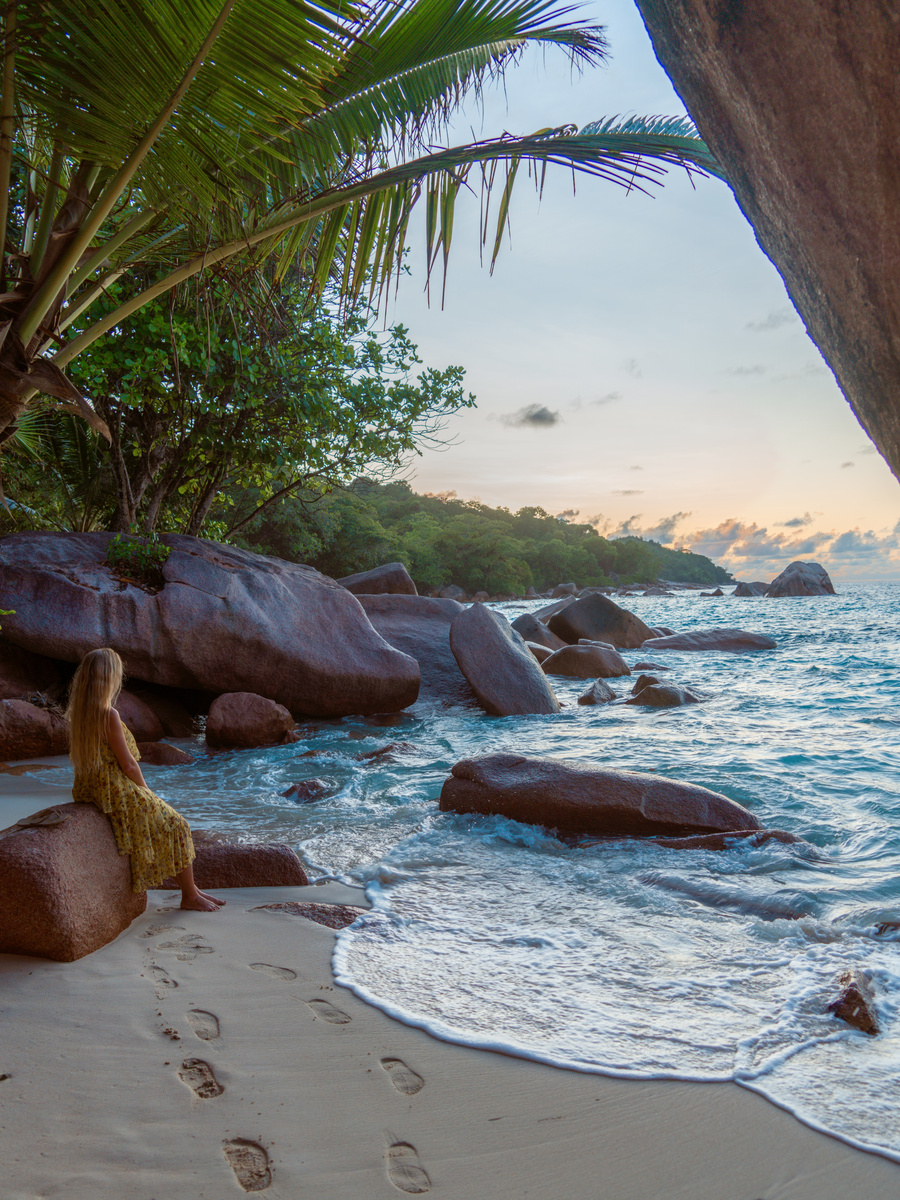 Olga sitting beside granite boulders as waves wash into a cove near Anse Lazio at dusk.