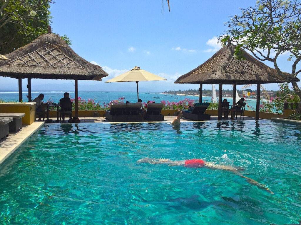 Guests swimming in Batu Karang’s pool beneath thatched pavilions overlooking Jungutbatu Bay.