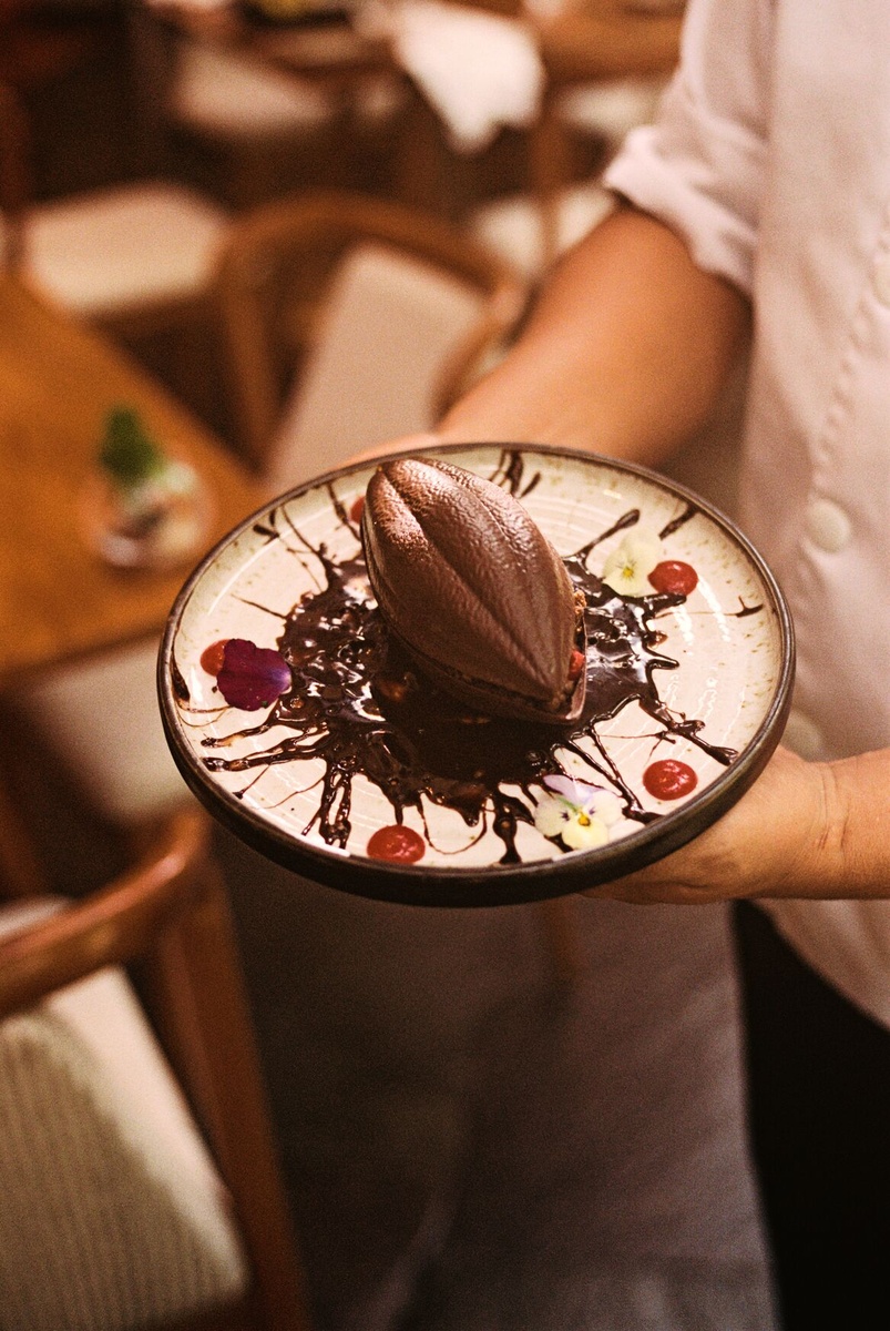 A server holds a plate with a dark dessert shaped like a cacao pod, garnished with flowers and sauce.