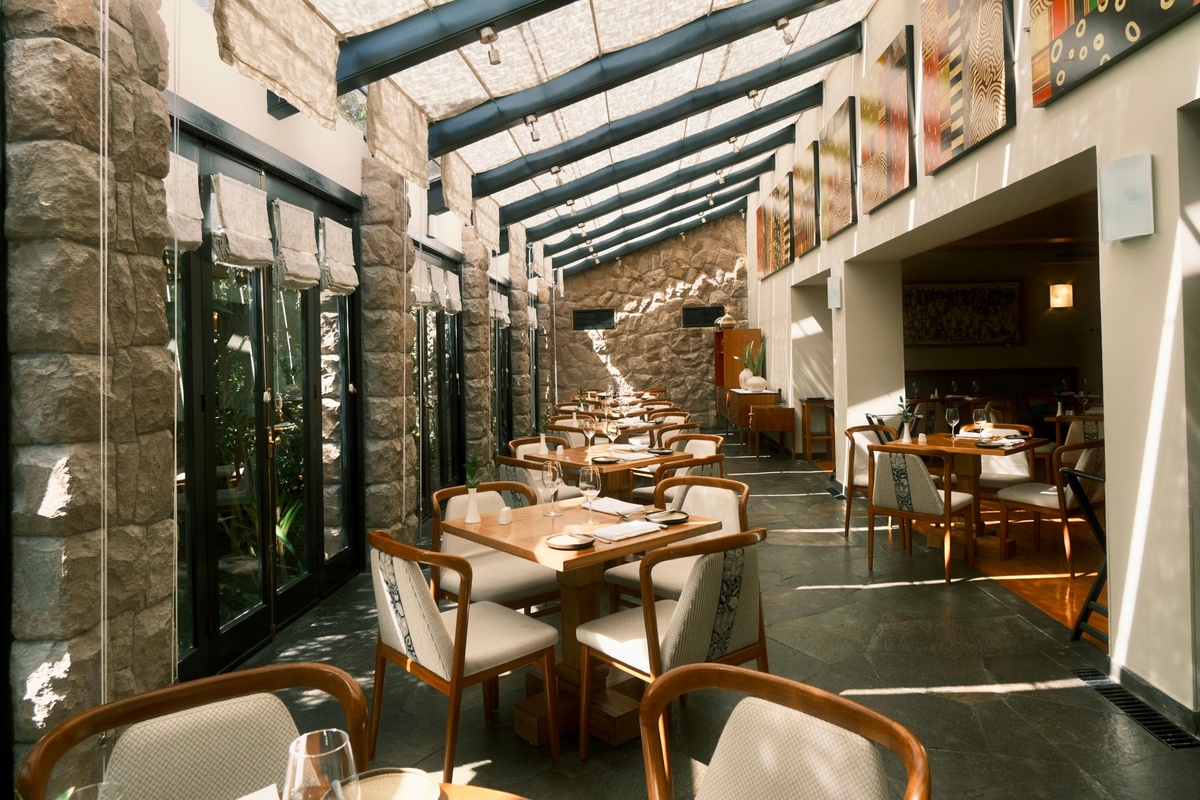 Set wooden tables stand beside tall windows and stone walls beneath Tampu Restaurant’s sloping glazed roof.