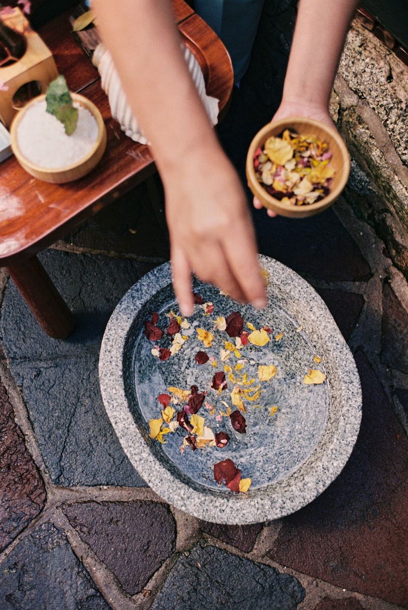 A therapist scatters petals into a stone basin beside bowls of treatment ingredients.