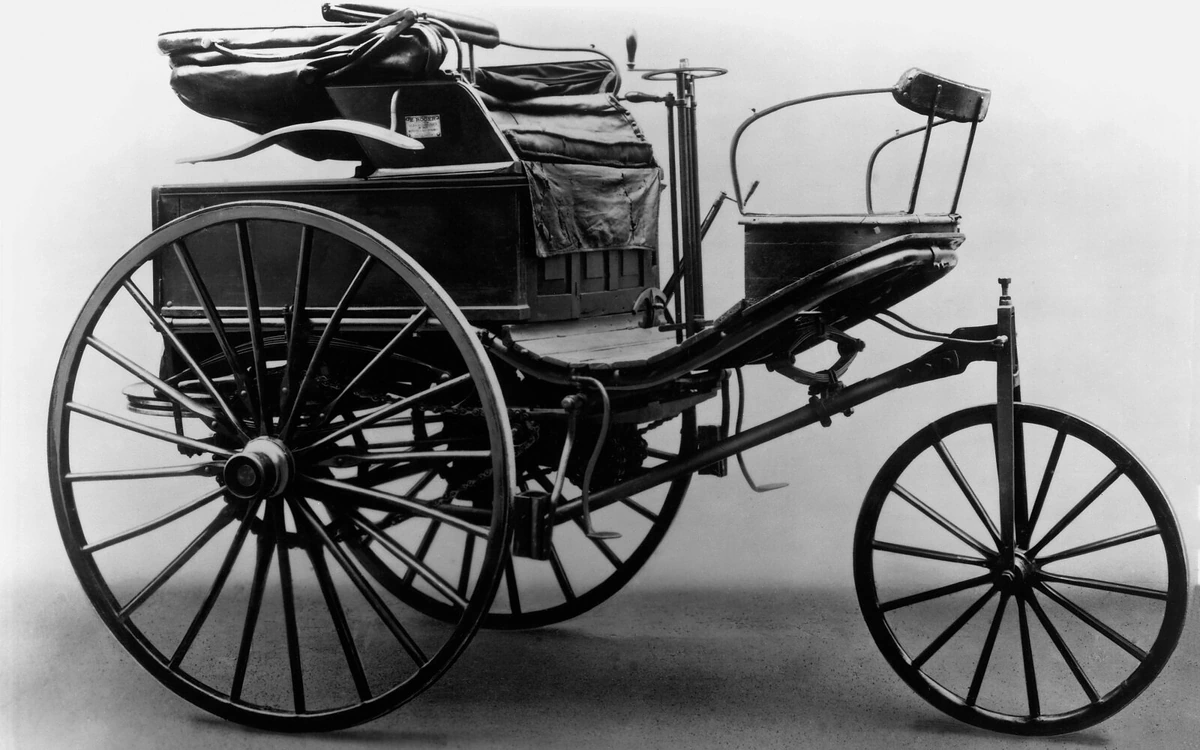 Black-and-white photograph of a Benz Patent-Motorwagen Model 3, with three wooden-spoked wheels, open seats and a folded hood.