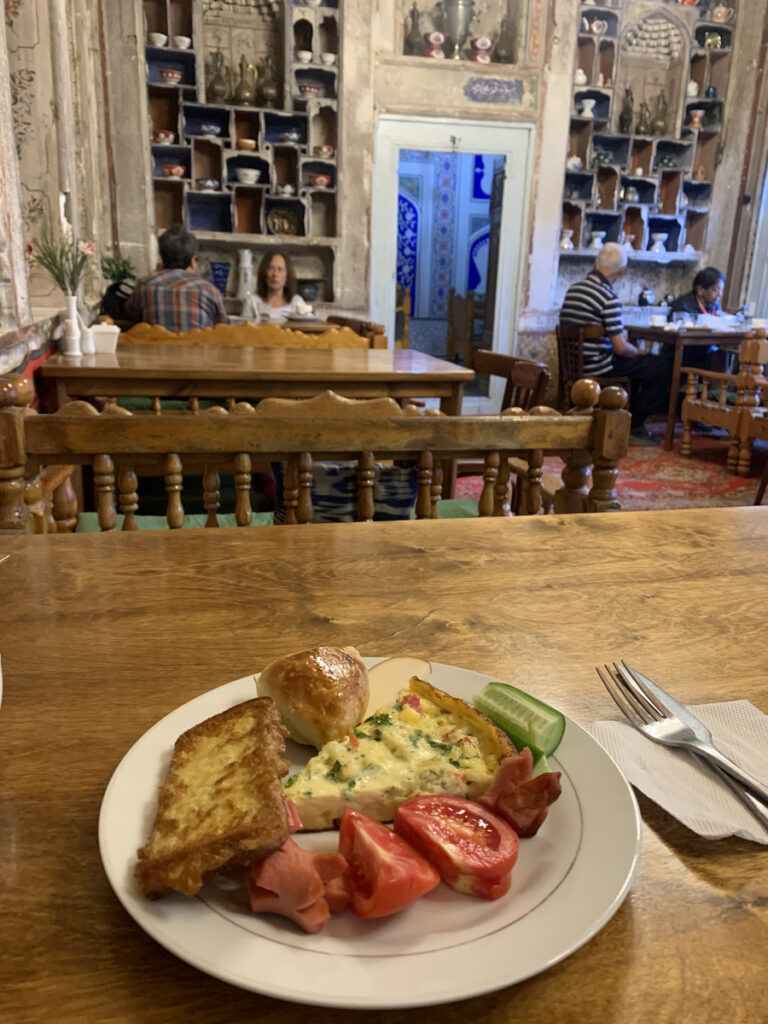 Breakfast on a wooden table in a historic Bukhara dining room lined with carved niches, ceramics and painted plaster.