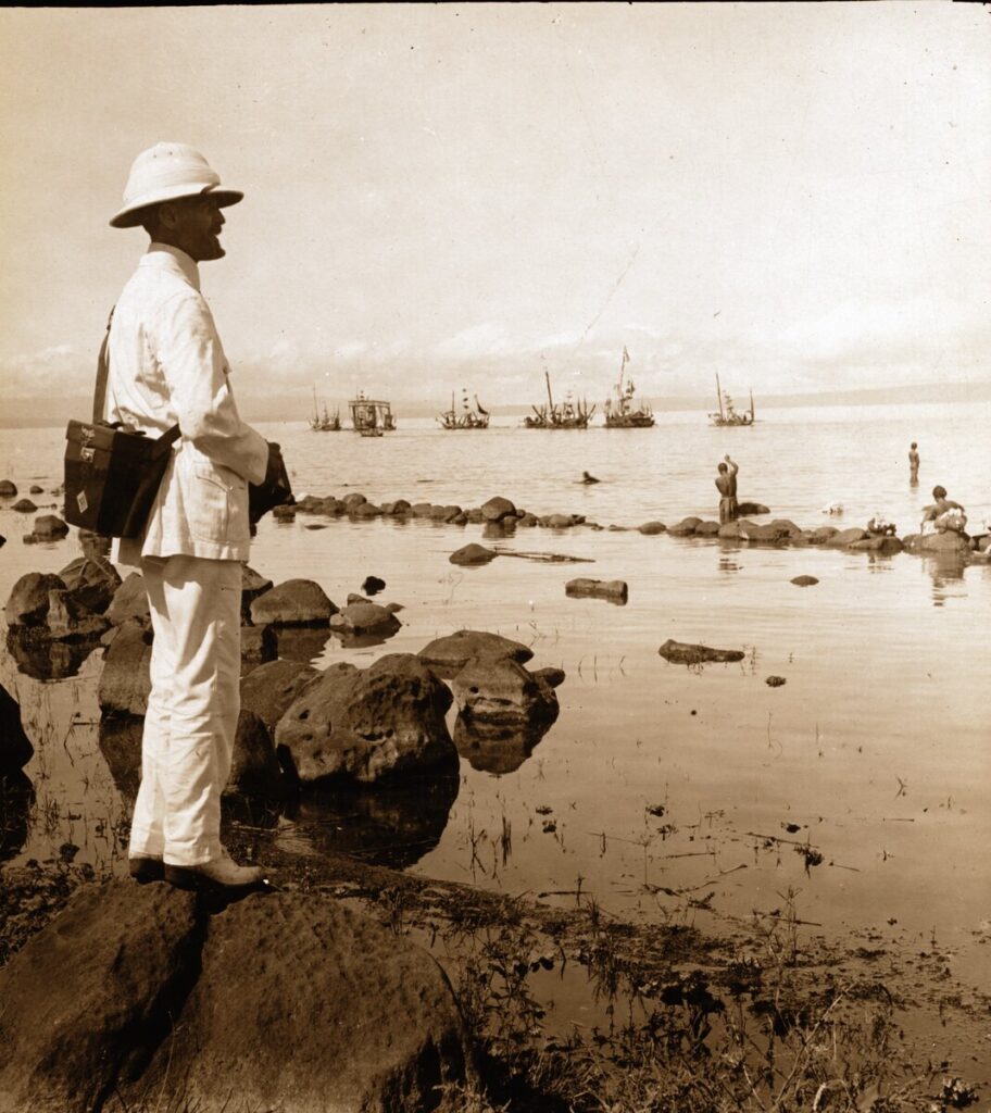Burton Holmes in a white suit and sun helmet stands on shore rocks with a camera case, looking across calm water at sailing boats.