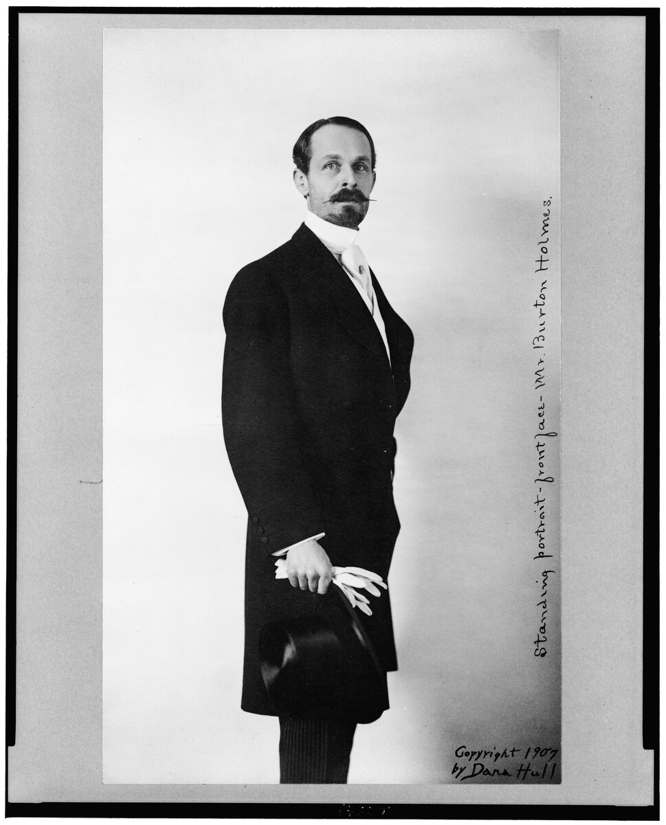 Full-length studio portrait of Burton Holmes in a frock coat, holding a top hat and gloves.