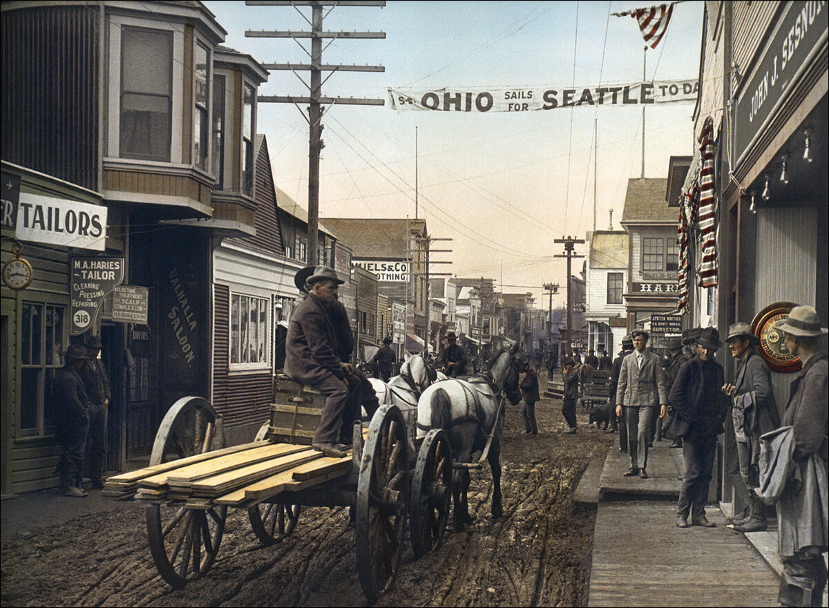 A hand-coloured view of a muddy street in Dawson City, with a horse-drawn cart of timber, wooden storefronts and a banner across the street.
