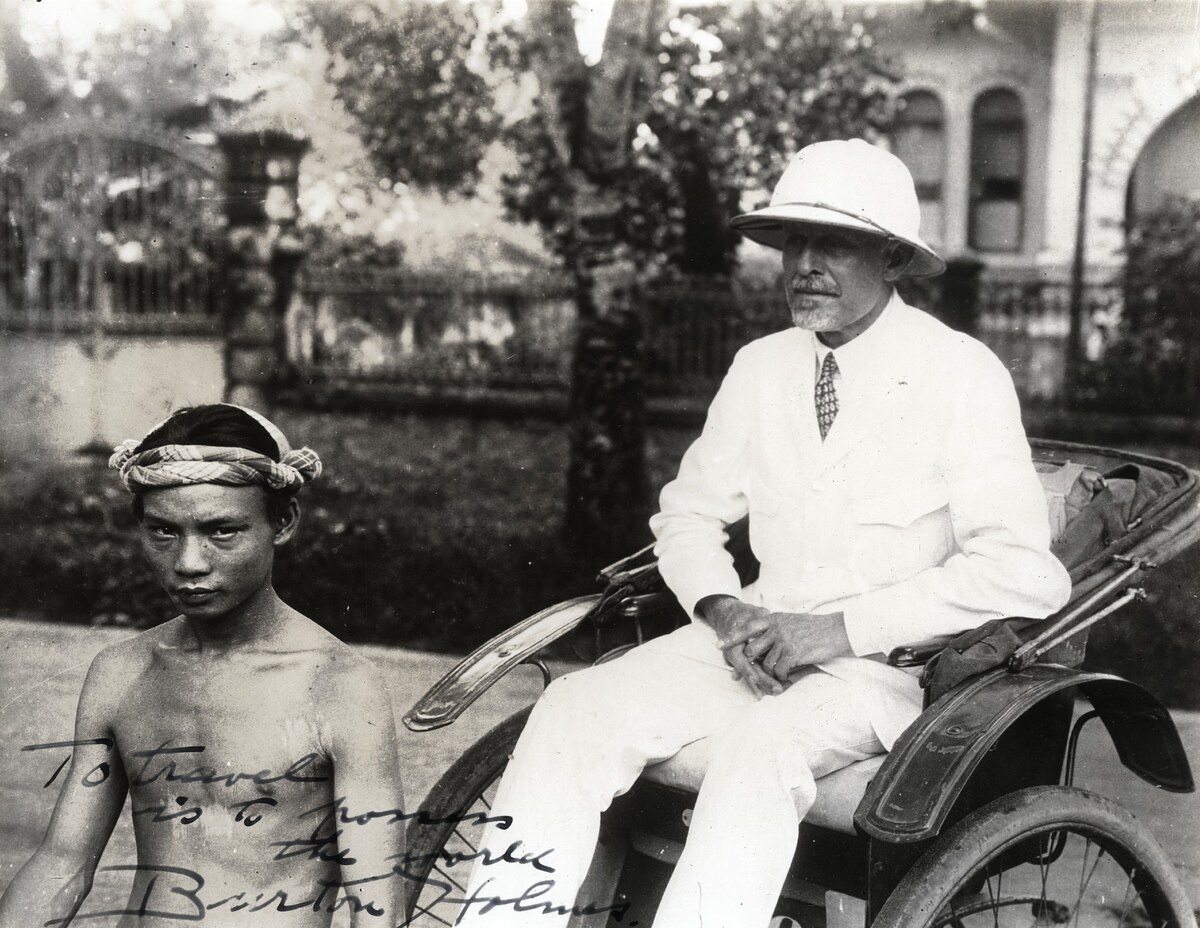 Burton Holmes in a white suit and sun helmet sits in a rickshaw beside a young bare-chested man who looks into the camera; a handwritten inscription is visible at lower left.