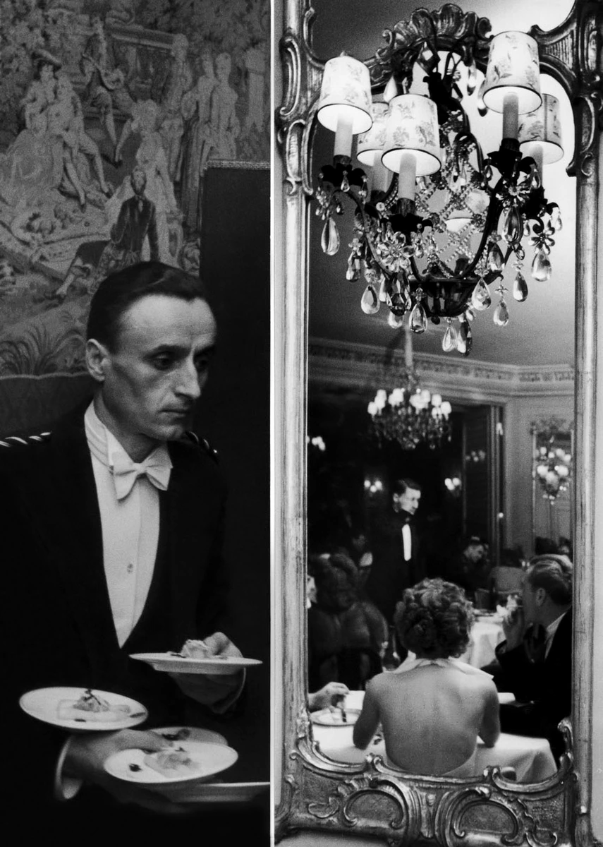 A waiter carries plates beside an ornate mirror reflecting formally dressed guests seated at dinner tables.