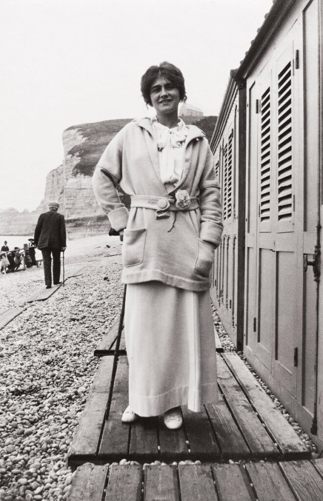 Gabrielle Chanel stands beside beach cabins at Étretat in a belted jersey jacket and long skirt, with her hands in her pockets.