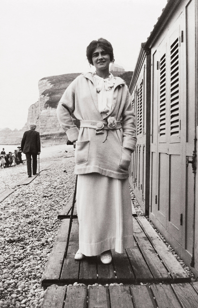 Gabrielle Chanel stands beside beach cabins at Étretat in a belted jersey jacket and long skirt, with her hands in her pockets.
