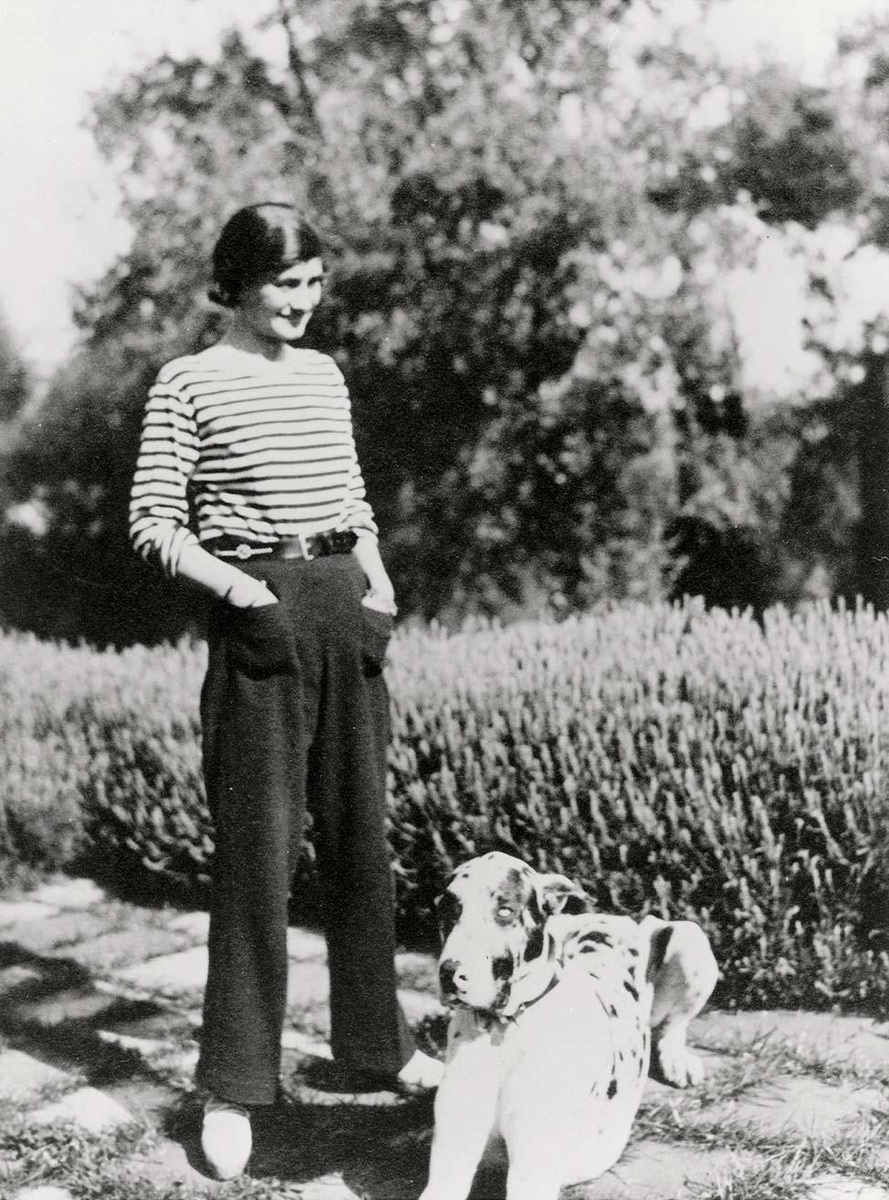 Gabrielle Chanel stands in a garden wearing a striped sailor’s shirt, wide-legged trousers and light shoes, with her hands in her pockets and her dog Gigot beside her.