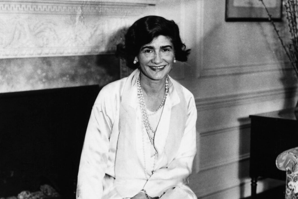 Gabrielle Chanel smiles in a white blouse and pearl necklaces beside a fireplace at the Hotel Pierre.