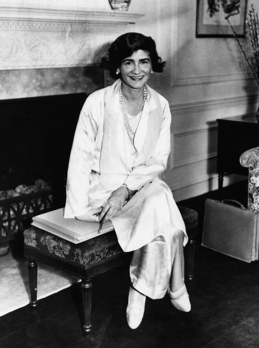 Chanel sits smiling in white satin trousers and an open-neck blouse with long pearl necklaces in a hotel room.