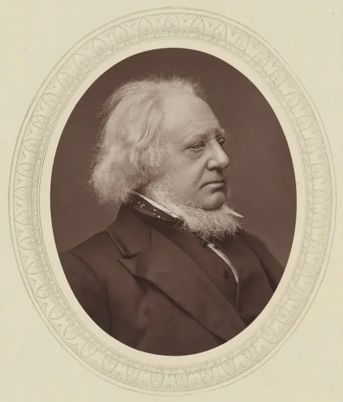 Henry Cole in profile, with wispy white hair, a white chin-fringe beard and a dark coat.