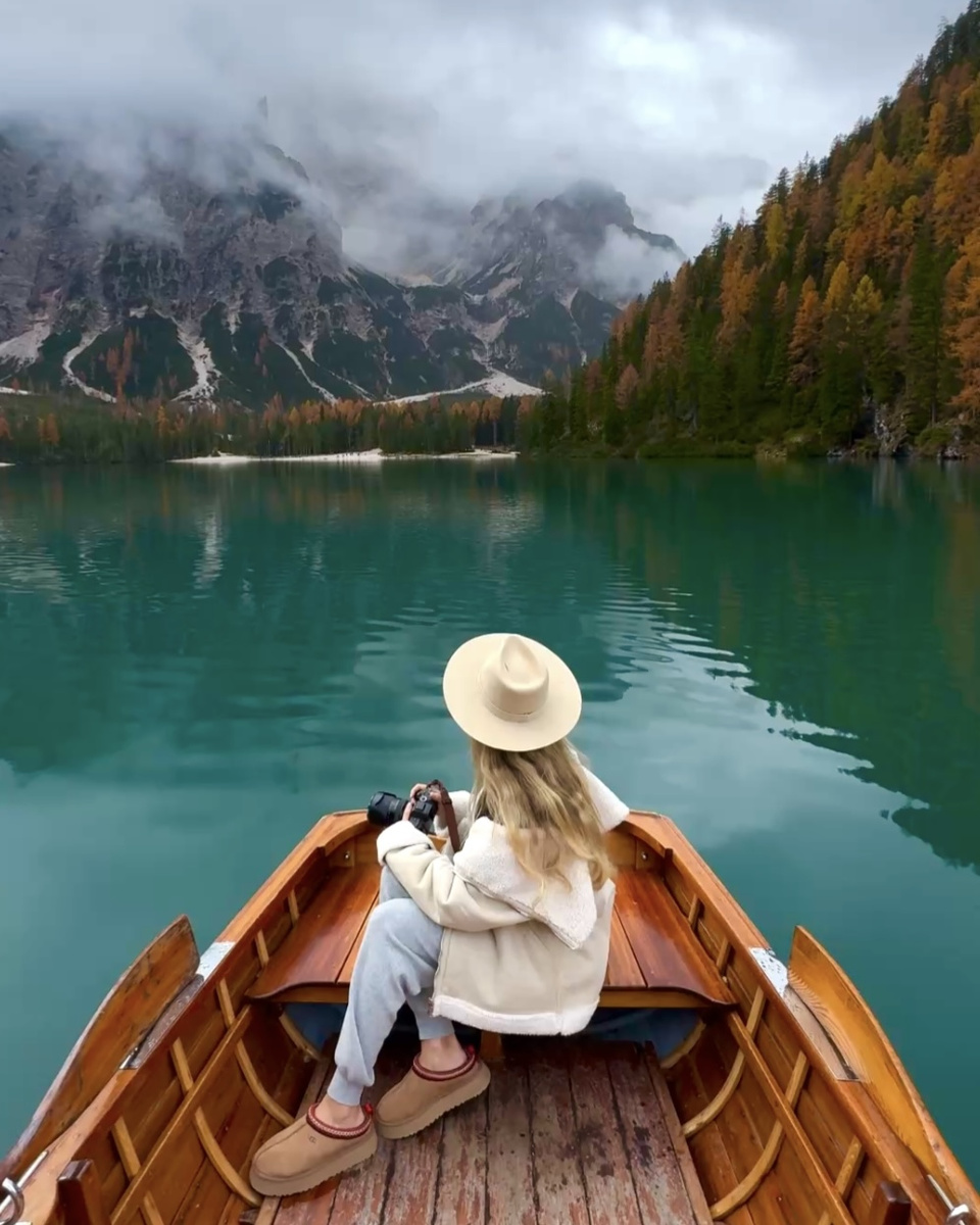 Olga holds her camera in a wooden rowboat on Lago di Braies beneath cloud-covered mountains.