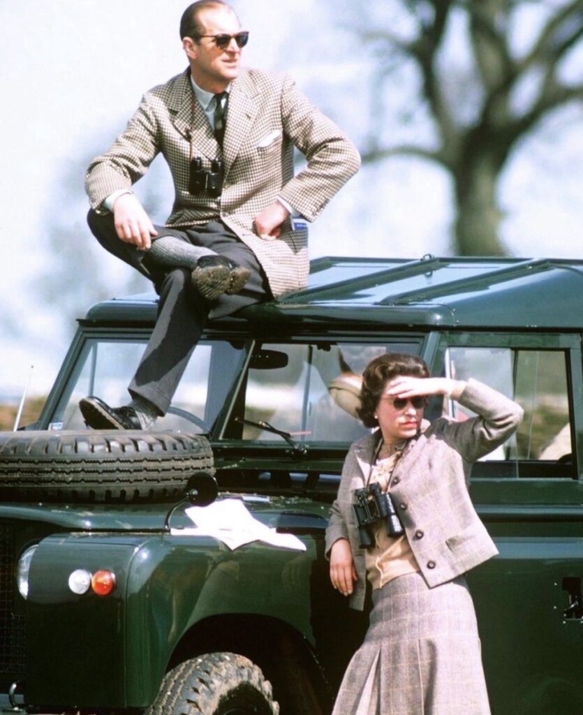 A man sits on the roof of a green Land Rover while a woman stands beside it; both wear binoculars, and she shades her eyes.