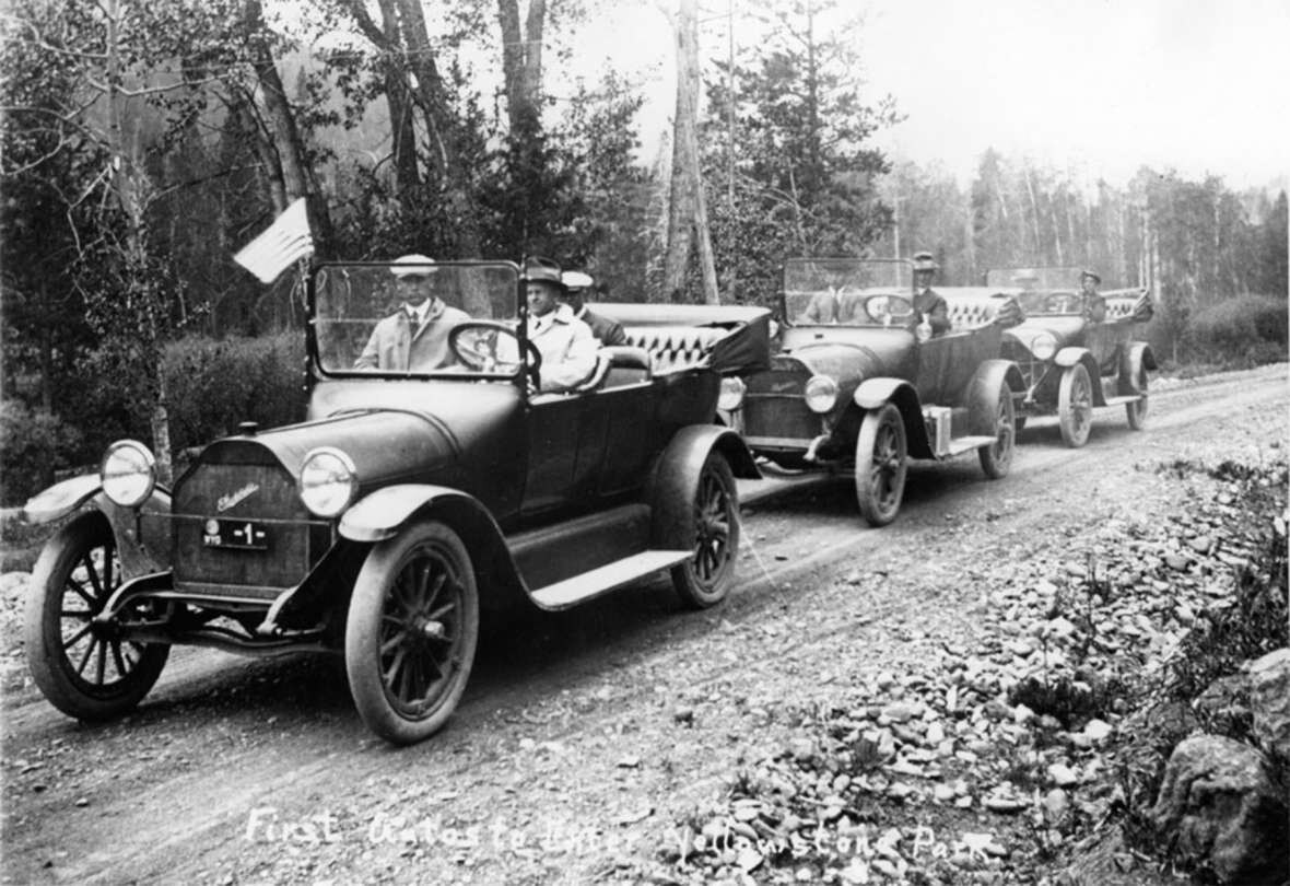 Three open touring cars with passengers on a gravel road through pines, an American flag flying from the lead car.