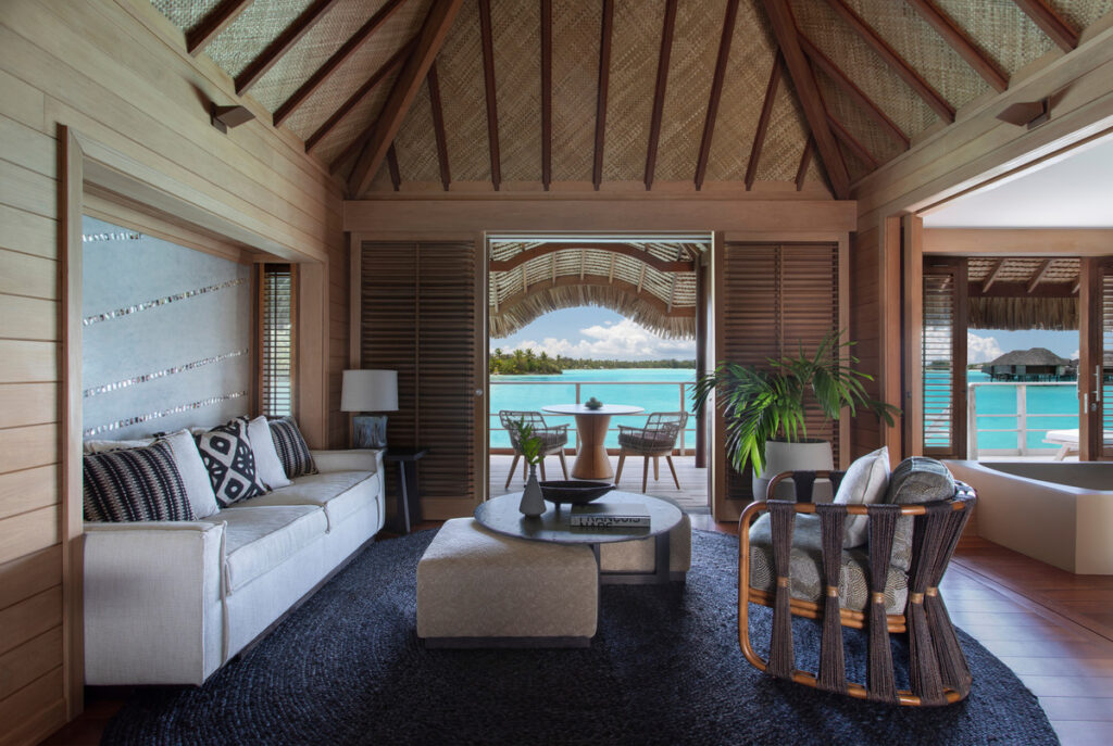 Teak-lined overwater bungalow living room with a sofa and open doors to a terrace above the Bora Bora lagoon