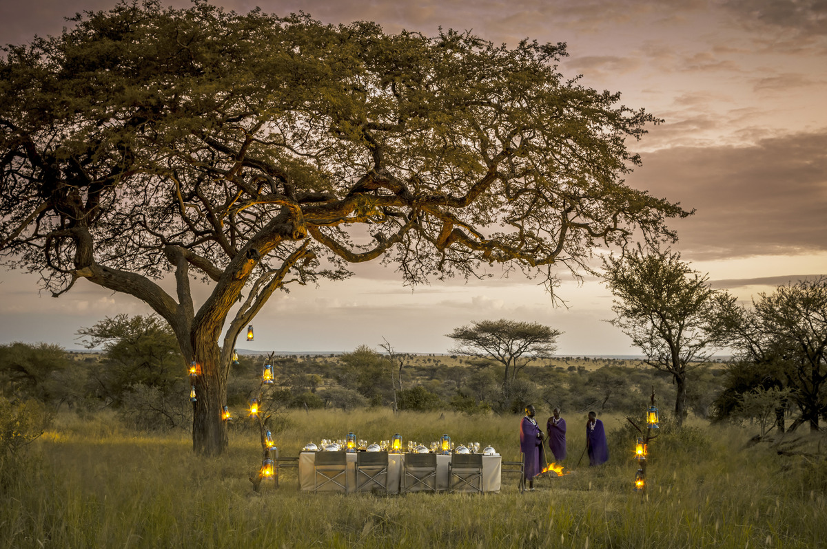 A white-clothed dinner table and canvas chairs beneath a lantern-lit tree, with a fire and three people beyond, in the Serengeti at dusk.