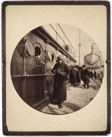 Circular photograph of George Eastman holding a box camera on a ship’s deck, with other passengers behind him.