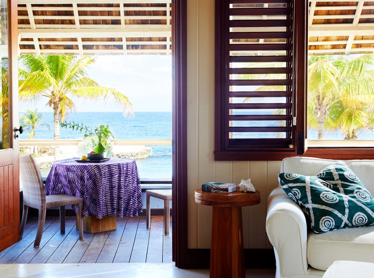 A beach villa’s white sofa and wooden shutters beside a veranda overlooking palms and the sea.
