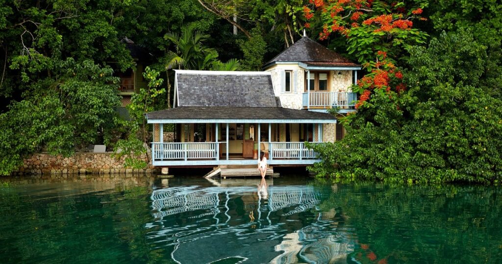 A white villa with blue railings beside the lagoon at GoldenEye, with a woman seated on its waterside steps.