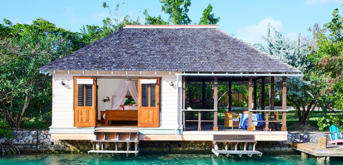 A white cottage with open wooden doors and a shaded veranda above turquoise water at GoldenEye. Courtesy of GoldenEye.