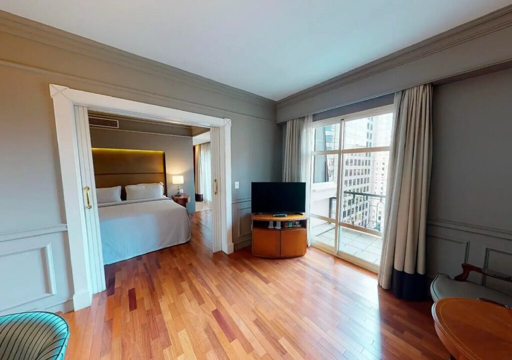 A Deluxe Suite, with the bedroom beyond a sitting area and tall windows opening to a balcony.