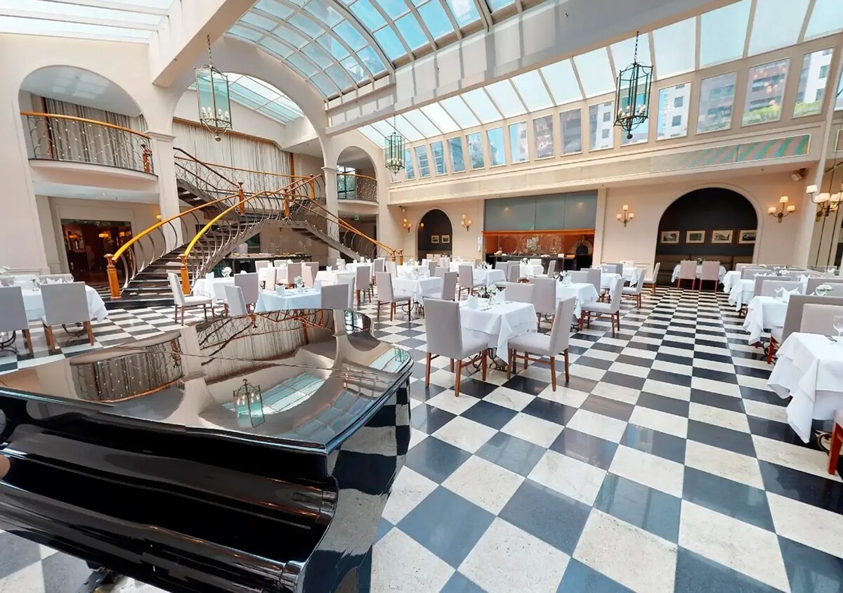 The Plaza restaurant under a glass roof, with a grand piano, a chequerboard floor and a curving double staircase.