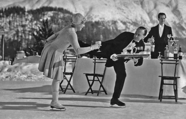 A woman on skates receives a drink from a waiter in formal dress beside tables on a snowy Alpine ice rink.