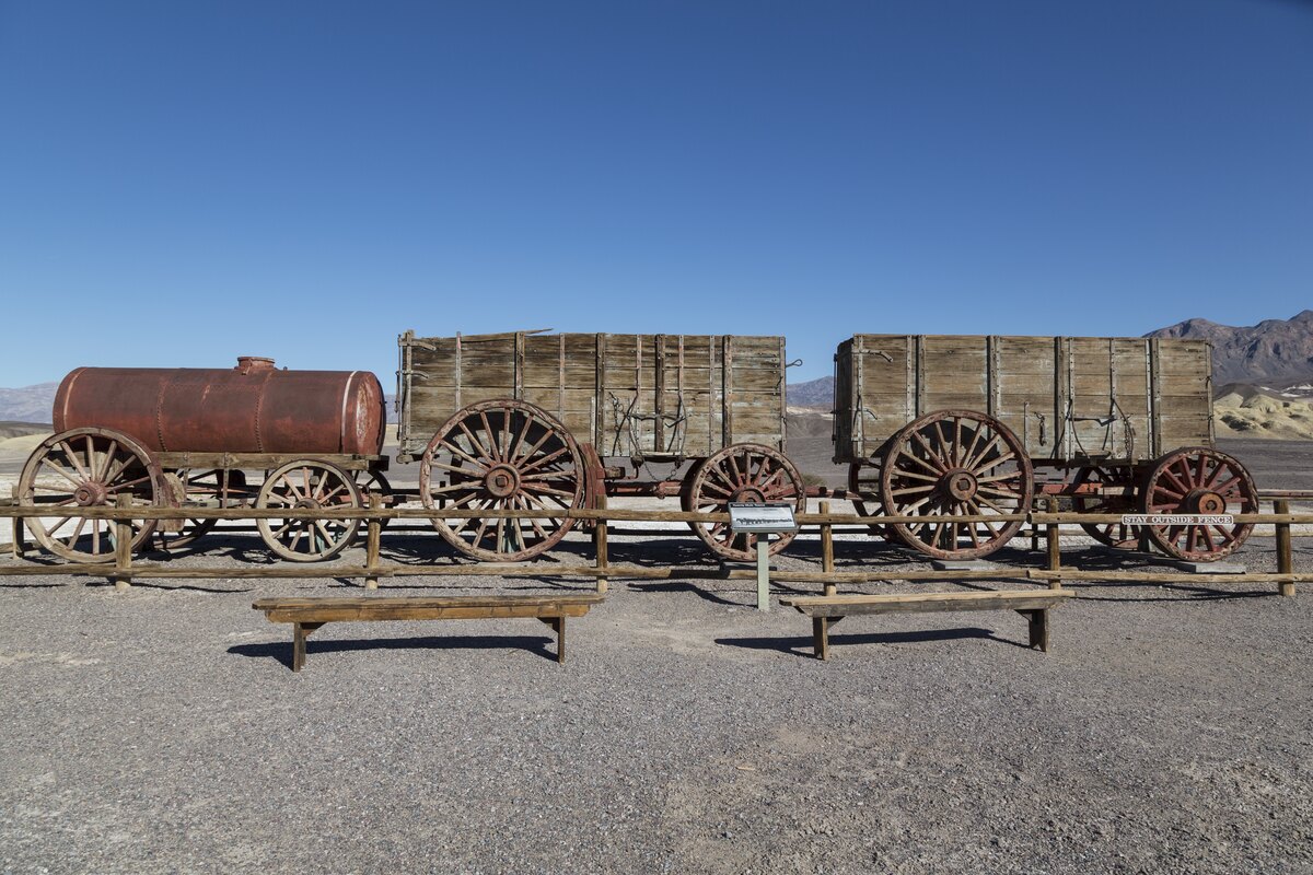 Two large wooden ore wagons and a rusted water-tank wagon stand behind a rail fence in bare desert under a blue sky.