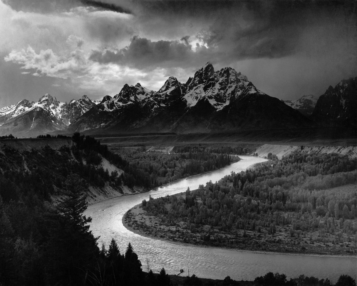 A black-and-white photograph of the Snake River winding through a dark valley towards the jagged, snow-streaked Teton range under a stormy sky.