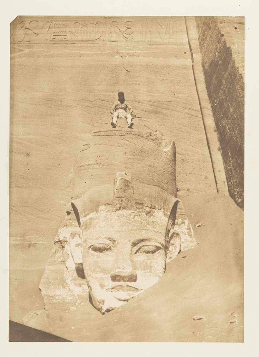 A sepia photograph of the enormous carved head of Ramesses II at Abu Simbel, half buried in sand, with a small man seated on the crown.