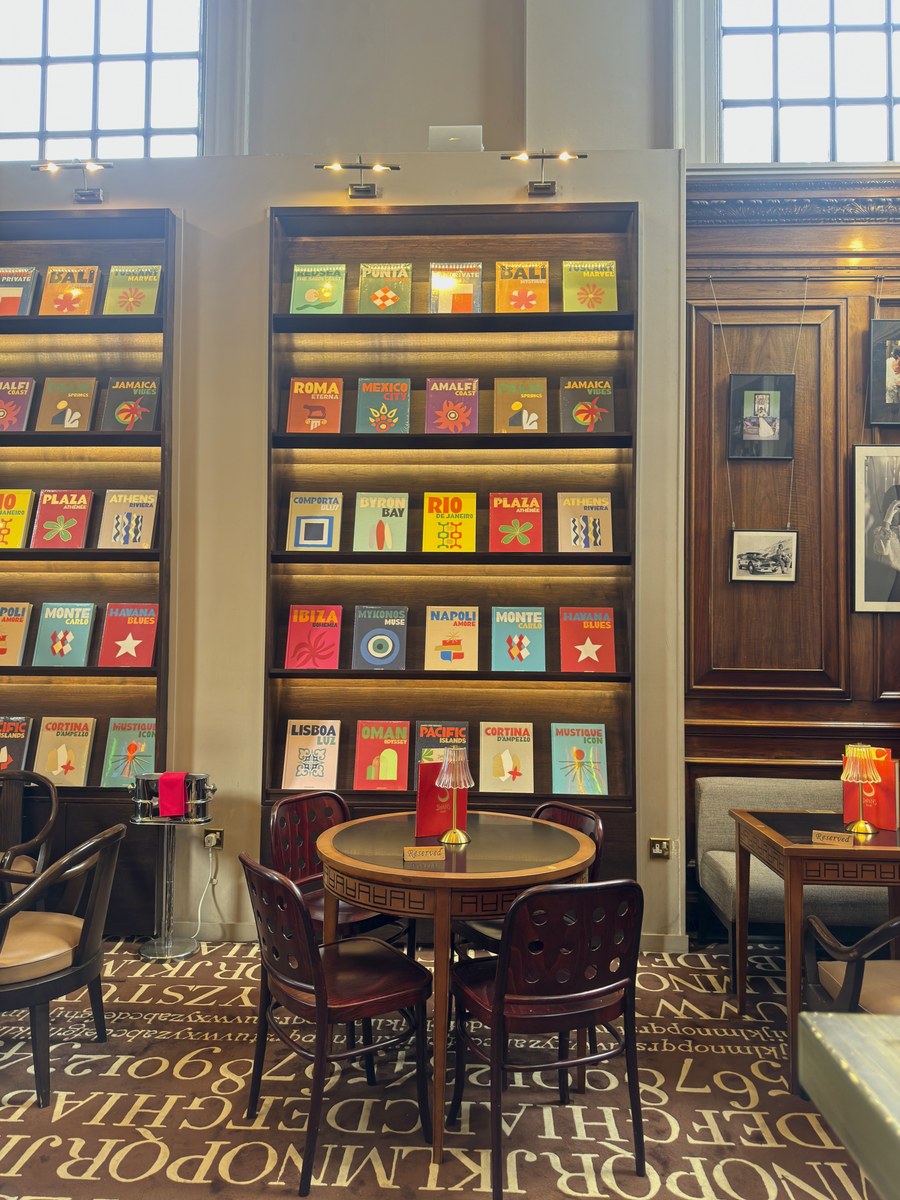 The interior of Maison Assouline in London, with tall illuminated shelves of books, dark wood panelling, a round café table and chairs.