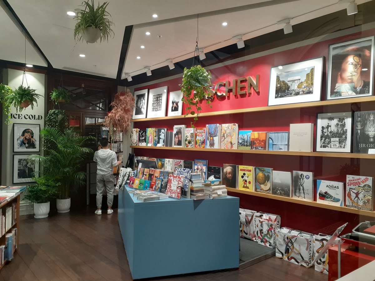 Shelves of large TASCHEN books with brightly coloured covers in a bookshop with red walls and potted palms.