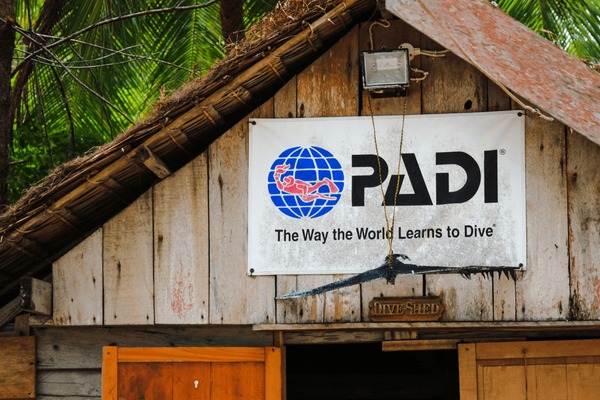 PADI sign on a weathered wooden dive hut with a rusty roof and a hanging fish silhouette.