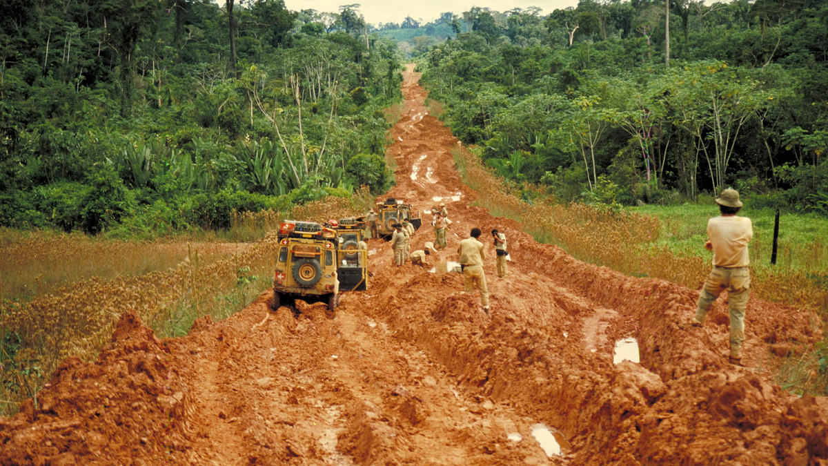 Yellow expedition vehicles and crews stand in deep red mud on a track running through rainforest.