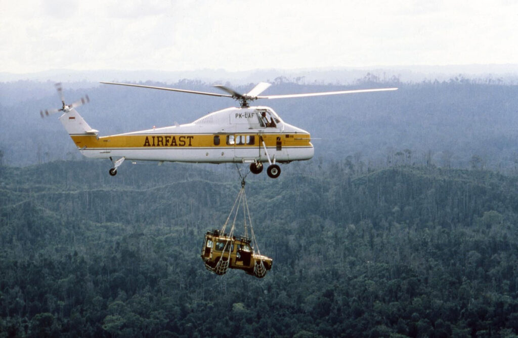 A yellow Land Rover hangs in a sling beneath an Airfast helicopter above rainforest.