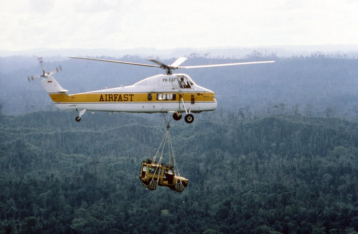 A yellow Land Rover hangs in a sling beneath an Airfast helicopter above rainforest.