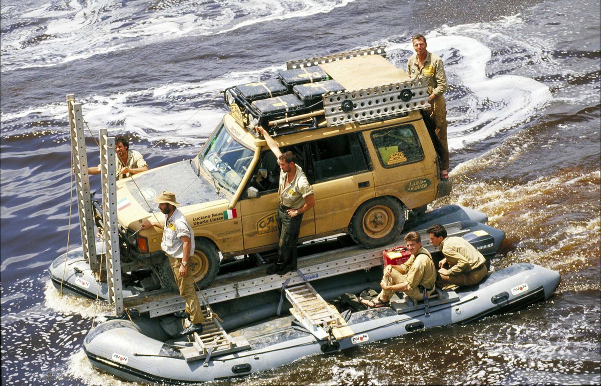 A yellow Land Rover Discovery and its crew cross a river on a platform supported by inflatable boats.