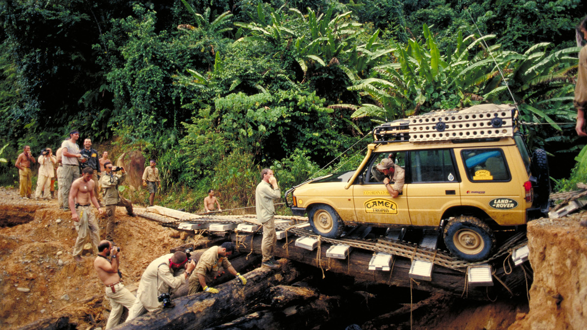 A yellow Land Rover Discovery crosses a timber bridge while people work around the vehicle in dense green forest.